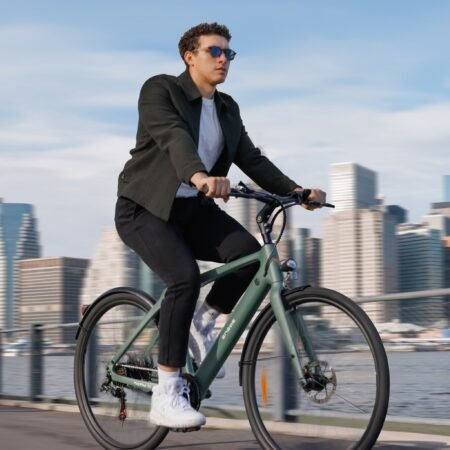 A person wearing sunglasses and a jacket rides a bicycle along a waterfront path, with a city skyline blurred in the background.