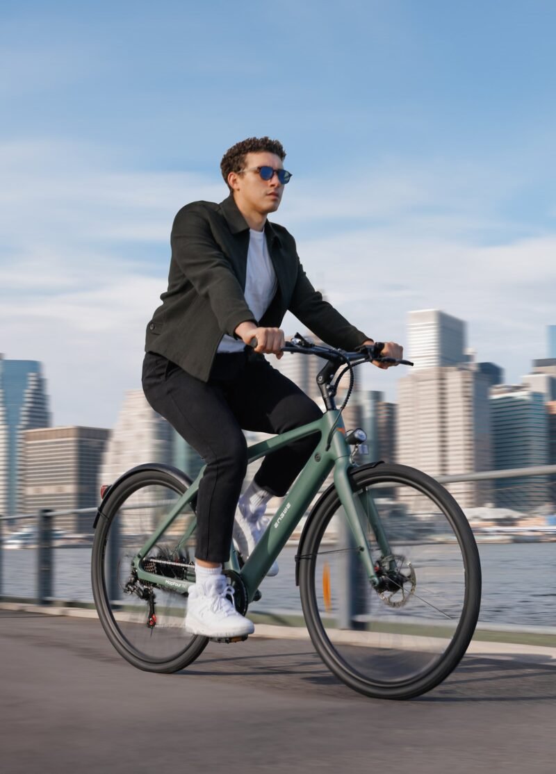 A person wearing sunglasses and a jacket rides a bicycle along a waterfront path, with a city skyline blurred in the background.